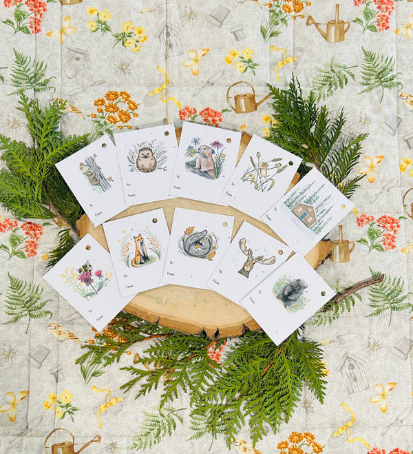 A 10 pack of plantable gift tags with hand drawn designs and 'to" and 'from' on the bottom. The designs are" bear cub in a tamarack tree, hedgehog, pika with flowers in its mouth, harvest mouse hanging from grain, a cabin in the woods, a bumblebee on wildflowers, a fox with autumn leaves are around him, a cozy squirrel, a moose with snow on his head, and a porcupine in flowers. 